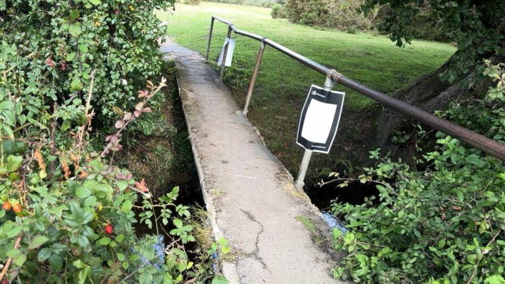 A narrow foot bridge crosses a stream with bramble on either side of the entrance. The bridge has a concrete layer that is cracked and a single metal handrail that is leaning to one side.