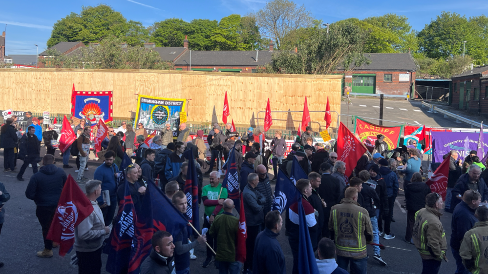 A large group of protestors gathered with large flags outside a bin depot. Some wear fire service uniforms of brown with high-vis markings. Behind them is a wooden fence with a gap in it for vehicles, which the people are blocking.