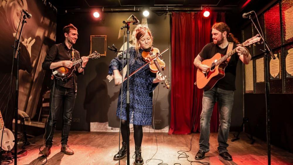 The Shackleton Trio, performing on a stage with coloured lights and decorations, playing instruments. Central in the image is Georgia, playing a violin, wearing a blue and black dress, with black leggings and boots. Her partner, Aaron Bennett is to the right, playing a guitar. He is dressed in a black top and faded black jeans. A third member of the band is on the left and he is wearing a brown shirt and black trousers, playing a banjo.