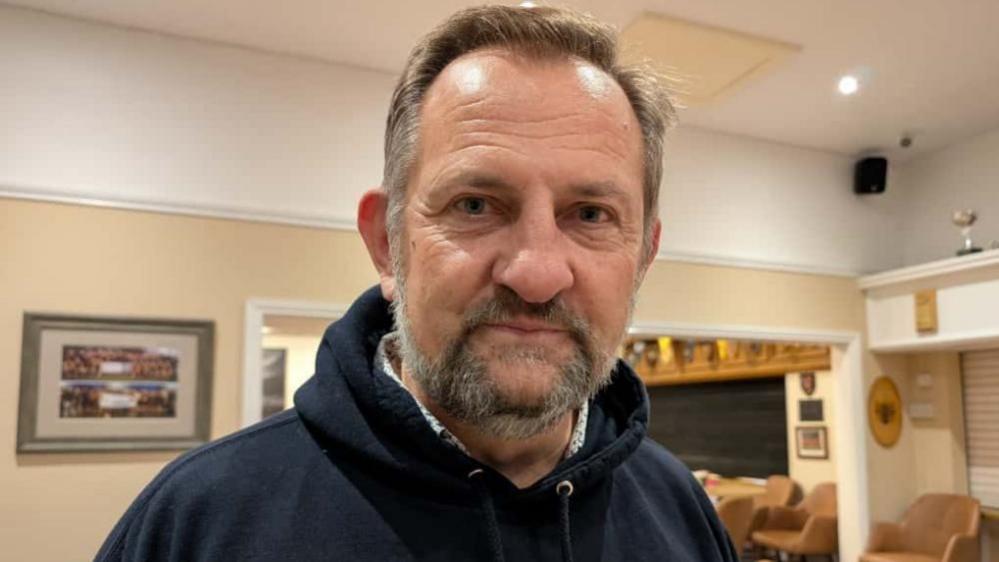 Founder Dan Wooler, a middle-aged man with a beard at Chard RFC clubhouse. He is wearing a dark navy hoodie with a shirt underneath.