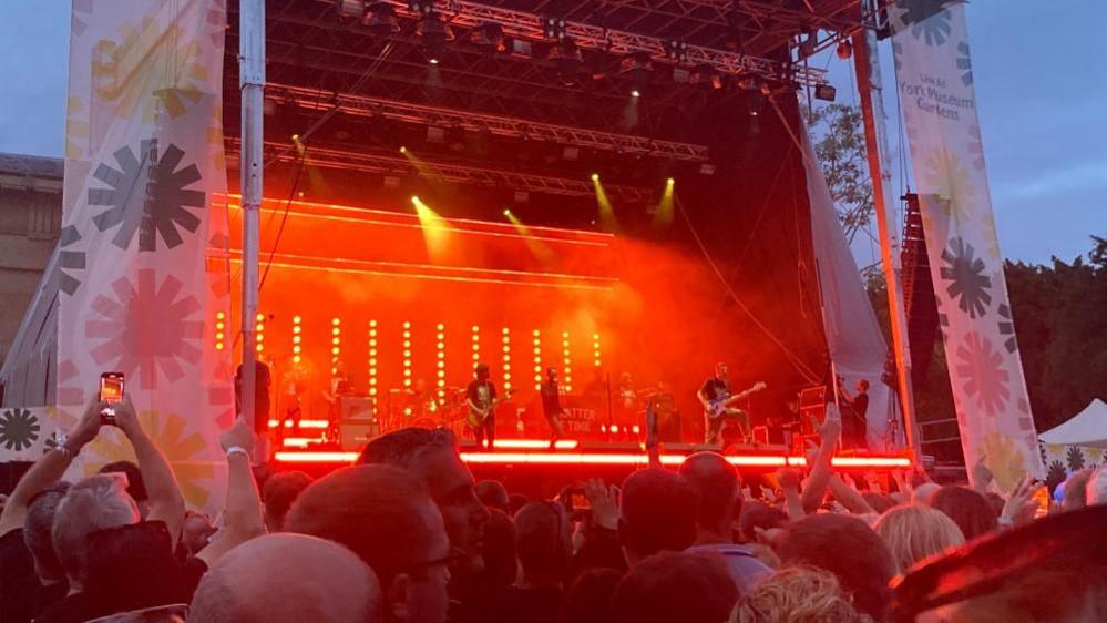 Shed Seven perform before a crowd, with banners adorning the stage reading "Live at York Museum Gardens".