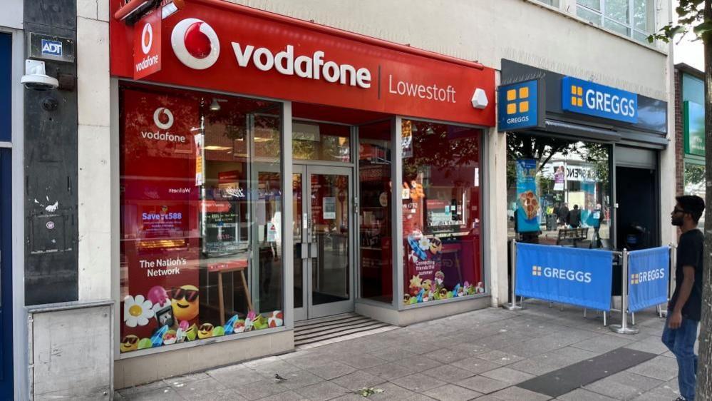 The Vodafone shop in Lowestoft, bearing the company and town name on the red signage, with text in white. The building next door is branded as Greggs bakery, with blue canvas fencing outside.