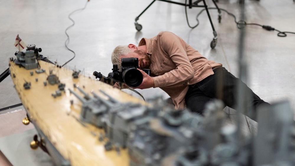 A photographer on the floor as he takes pictures of a model ship from several angles.