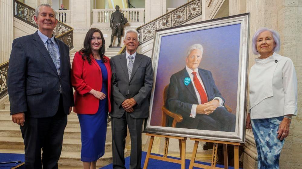 A group picture of Poots, left, is smiling at the camera. He is wearing a dark grey suit and light blue shirt. He has combed back grey hair. Little-Pengelly is next in the line. She is wearing a bright red blazer and blue knee length dress. She is smiling at the camera and has long brown hair. Peter Robinson is standing next to the portrait which is in a silver frame. In the portrait he is wearing a dark coloured suit and red tie, he is sitting down and has white hair. Standing next to it he is wearing a grey suit and tie and has short white hair. On the right, Iris Robinson is smiling beside the portrait. She is wearing a white top which is asymmetrical, and a blue skirt. She has slightly purple short hair.