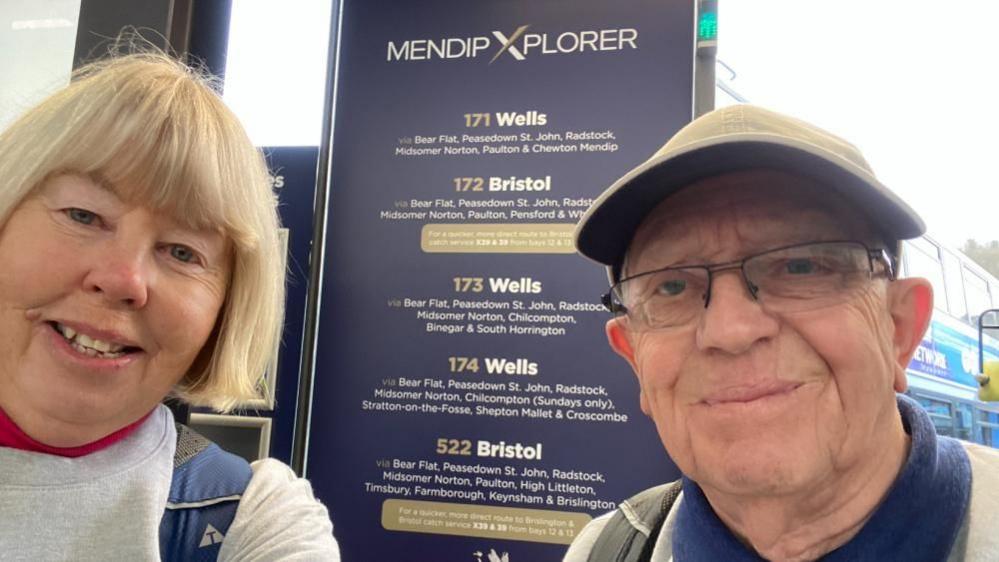 Mrs Banham, with blonde hair and wearing a light-coloured top with a blue backpack, and Mr Banham, wearing a light-coloured cap. They are standing in front of a notice showing bus routes in Somerset.