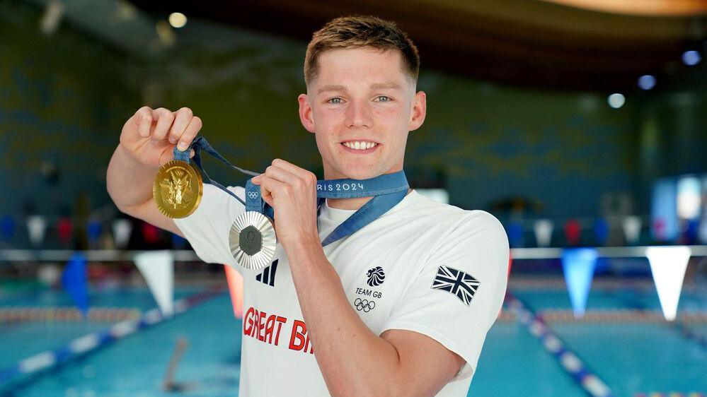 Duncan Scott smiles at the camera while holding up a silver medal and a gold medal which are both draped on ribbons around his neck. He stands in front of a swimming pool with red, white and blue bunting and wears a white Team GB T-shirt.