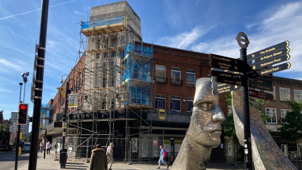 The outside of the former Debenhams store in Bedford with scaffolding up and shoppers walking past, next to a statue of two large metal faces 