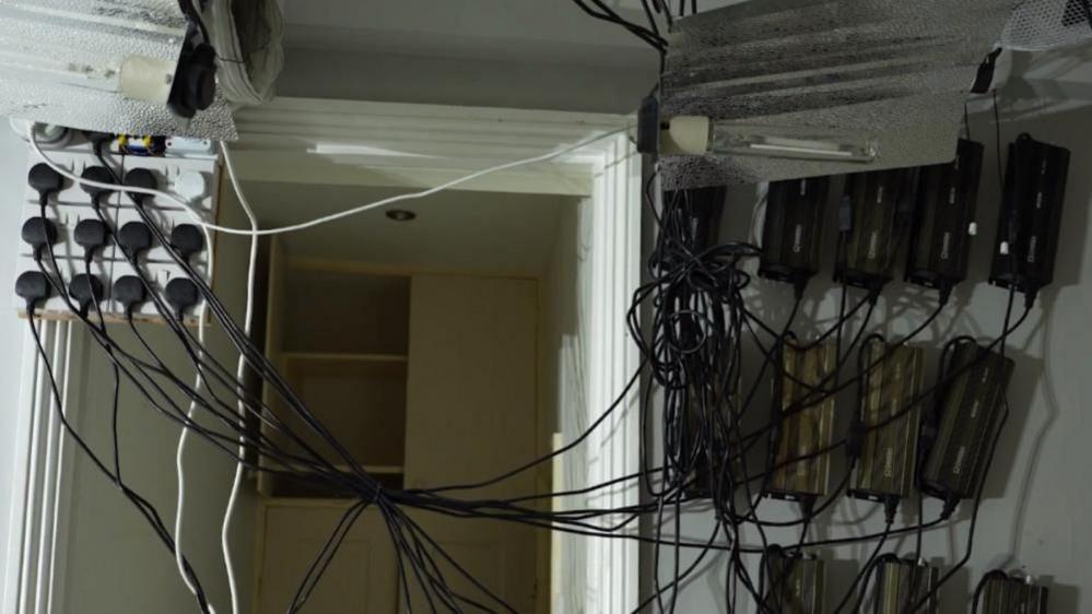 About 16 plugs, mostly black, are connected to sockets to the upper right of the picture. Black wires from the plugs stretch across the picture to black devices screwed into the wall. A white door frame is visible behind the wires, with a hole through which some of the wires have been installed.