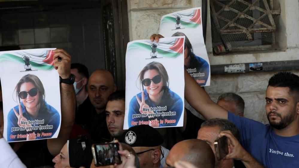 There is a crowd of people who hold up posters of a young woman with sunglasses and brown hair who is smiling and making a peace sign with her left hand. The poster says "May her blood pave the way for freedom. Our friend Stay in Peace."