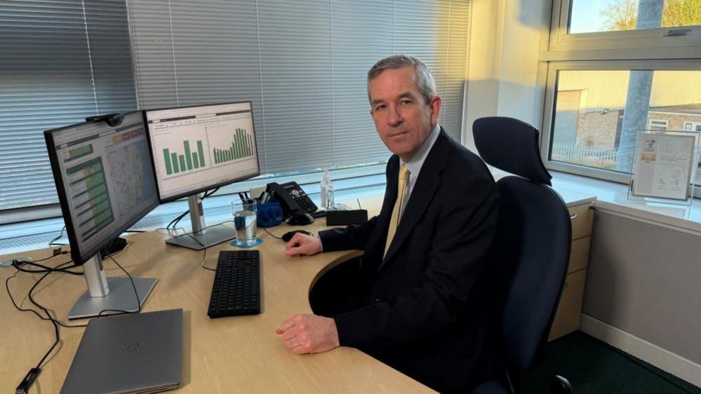 Neill Moloney with short grey hair looking at the camera wearing a dark suit and yellow tie. He is sitting at a wooden desk where there is a keyboard, a laptop and two monitors on stands.