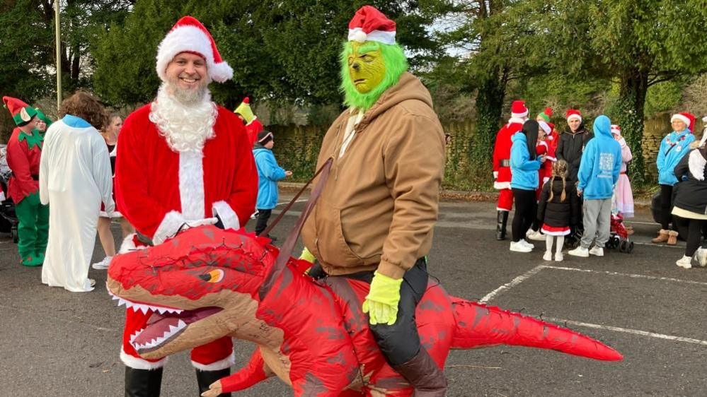 A man wearing a santa outfit, smiling at the camera, stood next to a man dressed as the Grinch, with a green mask and green fur on his face, who is riding an inflatable dinosaur