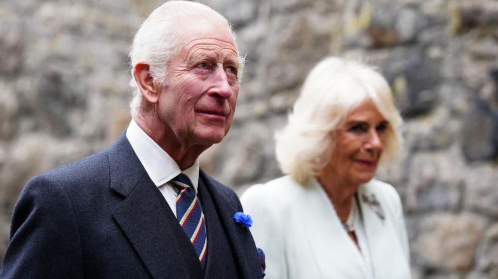 King Charles and Queen Camilla walking with a stone wall in the background. He is wearing a suit with a blue striped tie. She is wearing a white dress.