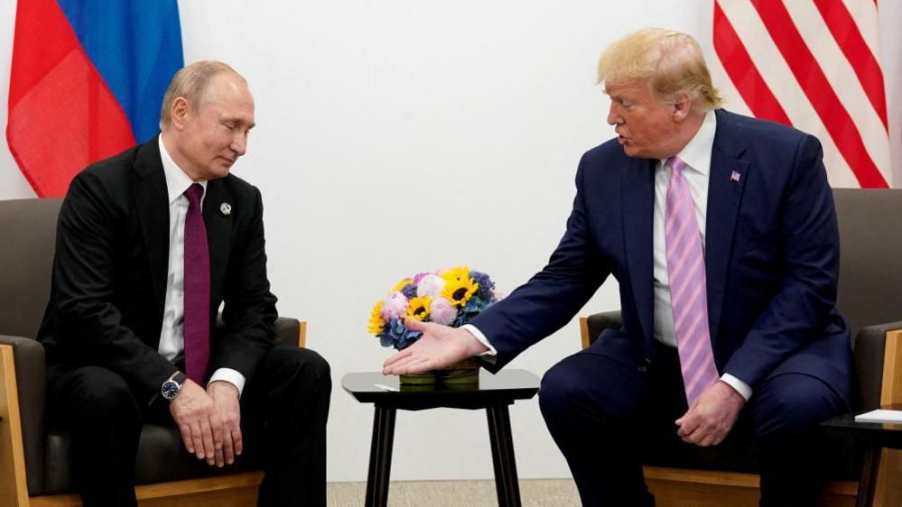 Russian leader Vladimir Putin, to the left of the picture, looks down at the hand of Donald Trump, who is speaking to him on the right. 

Both men are sat, wearing suits, with flags behind them. There are some flowers on the table between them. 
