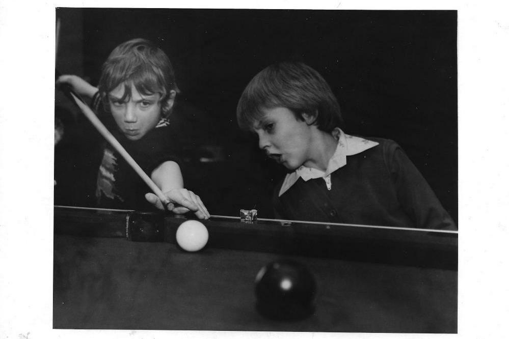 Two boys playing pool. The one on the left is using the pool cue and is pointing it towards the white ball with pursed lips. The other is looking at the ball from the side with his mouth open in a shocked expression.