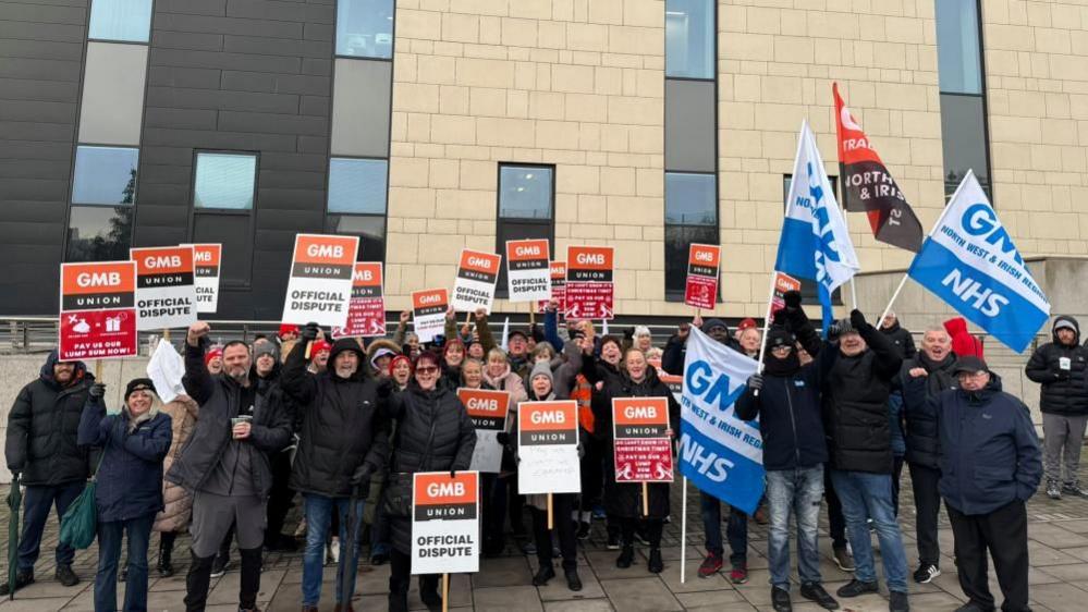 A large group of people stand outside a brick building holding signs and flads. Some have their fists in the air. 