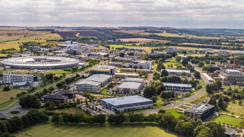 Harwell campus from the air. It is surrounded by ground fields, and there are tens of large buildings dotted around. The largest is a silver donut shaped structure.