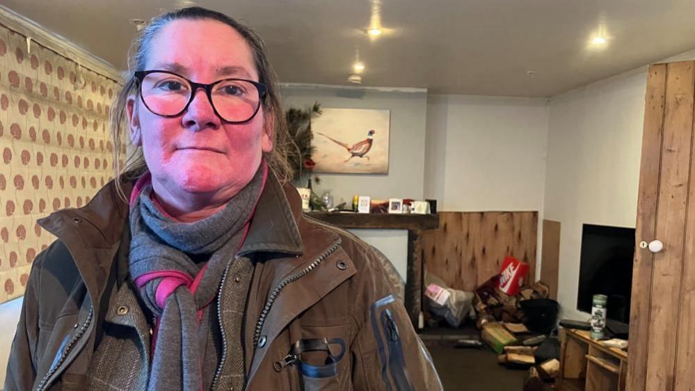 A woman wears glasses and wears a brown jacket while standing in a house with logs of wood on the floor