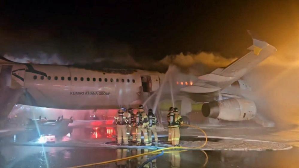 Firefighters tackle a blaze on board an Air Busan aircraft at Gimhae International Airport, South Korea. Picture shows the charred fuselage on the runway with smoke pouring out of it while firefighters standing close to the plane's tail spray jets of water on it.