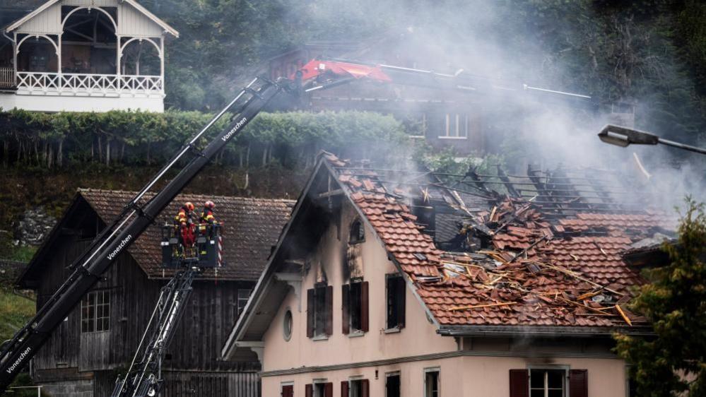 The building as pictured from the other side. Firefighters assess the damage from a cherry picker. About half of the riles on the roof are missing, with the rest burnt in the fire.
