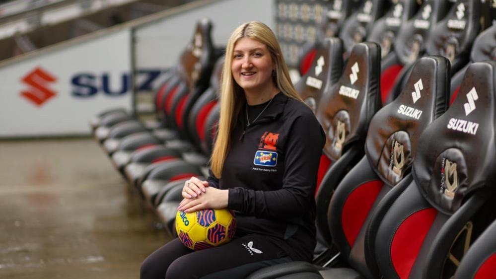 A woman with long, straight blonde hair and wearing a black tracksuit sitting in a dug-out at the MK Dons ground. She holding a yellow football on her knee. Empty red-and-grey seats are around her.