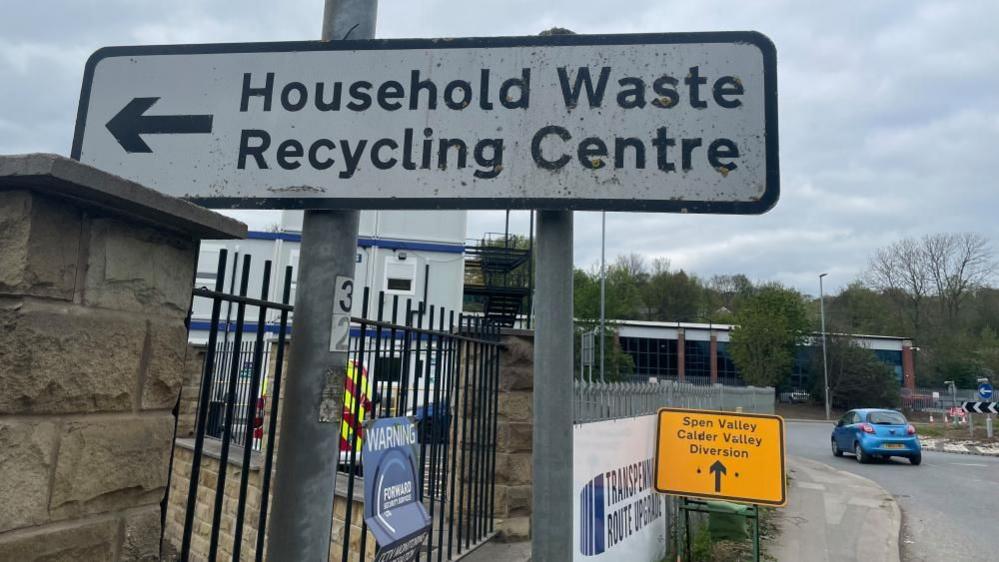 A sign with an arrow, followed by the words "Household Waste Recycling Centre". Another sign in the background reads "Spen Valley Calder Valley Diversion" and another says "Transpennine Route Upgrade". A car is going around a roundabout in the background.