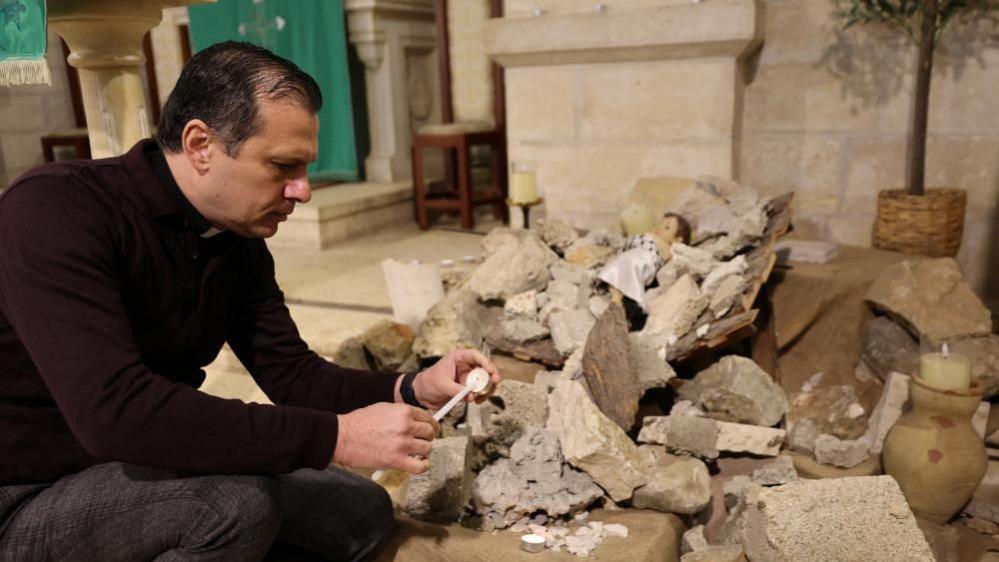 Reverend Munther Isaac lights a candle near a pile of rubble with baby Jesus figure lying on top inside the Evangelical Lutheran Church in Bethleham