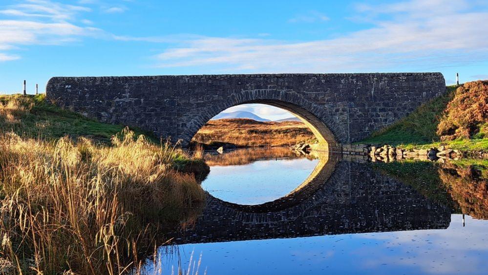 A flat, little stone bridge over a waterway reflects onto the water. It is surrounded by brown shrubbery with green, grassy banks.