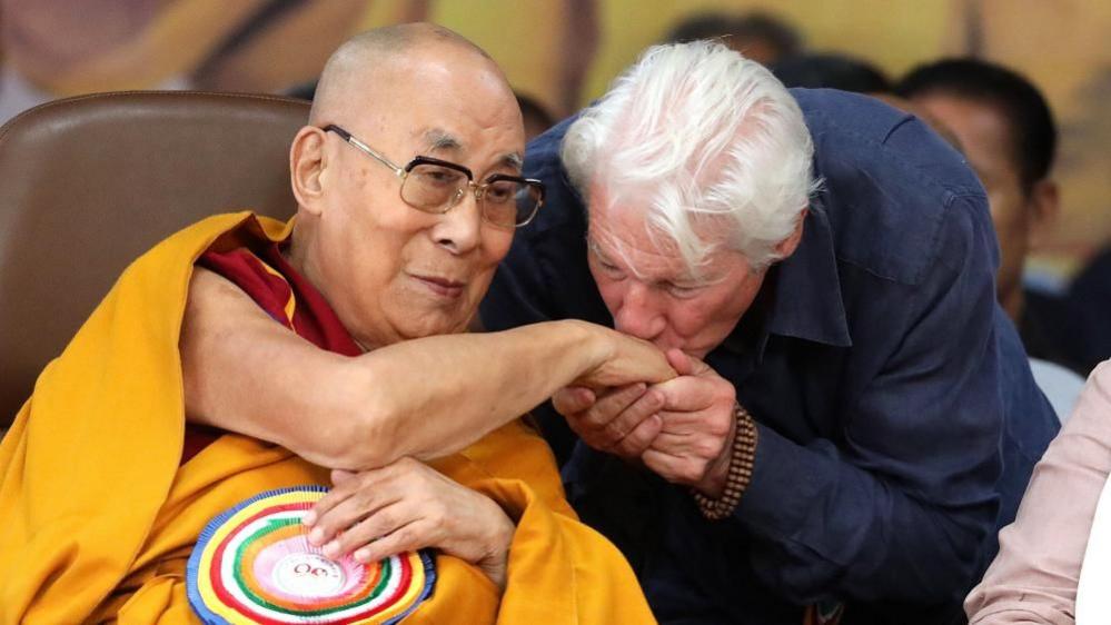 US actor Richard Gere, in a navy shirt and with wooden bracelets on, kisses the hand of the Dalai Lama cloaked in a yellow robe with a bright multi-coloured brooch on.