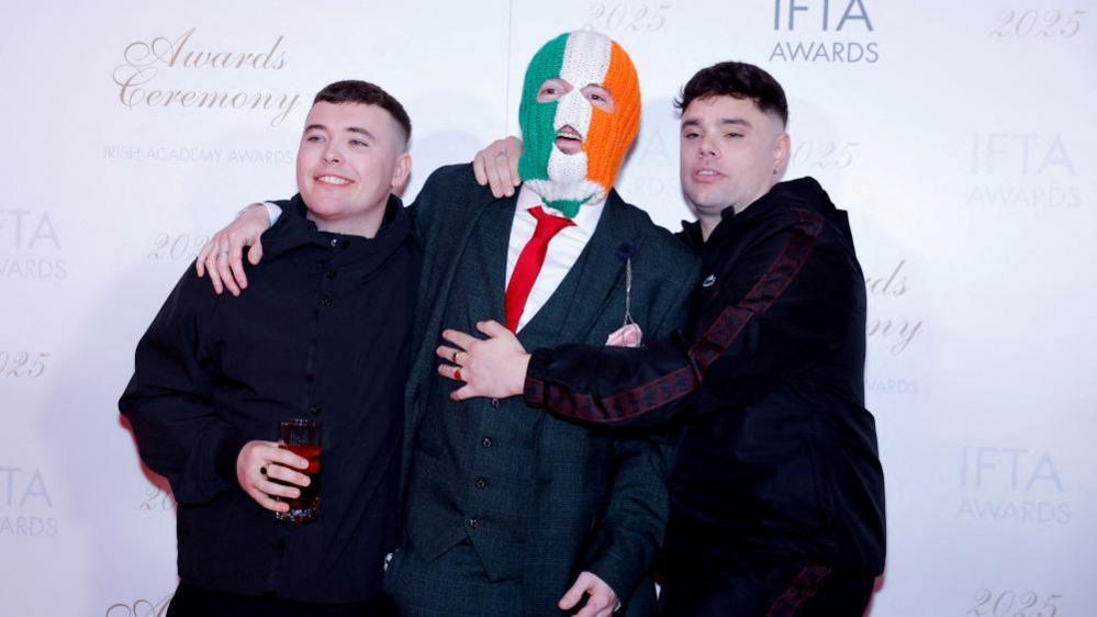 Three men are standing smiling for a photo. Left, is wearing a black coat and holding a glass. He is smiling at the camera and has short brown hair. The one in the middle is wearing a grey tartan suit and a red tie. He is also wearing a balaclava which is green white and orange striped. On the right, a man has his arms around the one in the middle. He is wearing a dark coloured jacket and has short black hair.