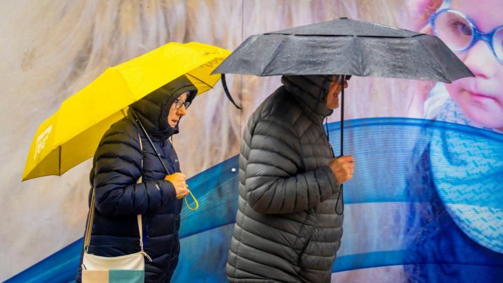 A man and a woman walk through a city centre past a billboard poster. The woman has a yellow umbrella and the man a black one. Both are wearing winter coats with the hoods up.