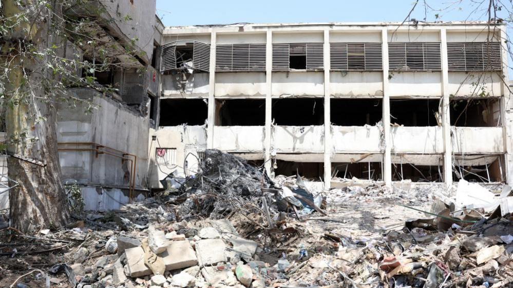 A white building with blown out windows is seen after an Israeli strikes on Evin prison in Iran. There is rubble piled high in front of the building, with piles of metal and concrete seen. In the foreground is a tree branch.