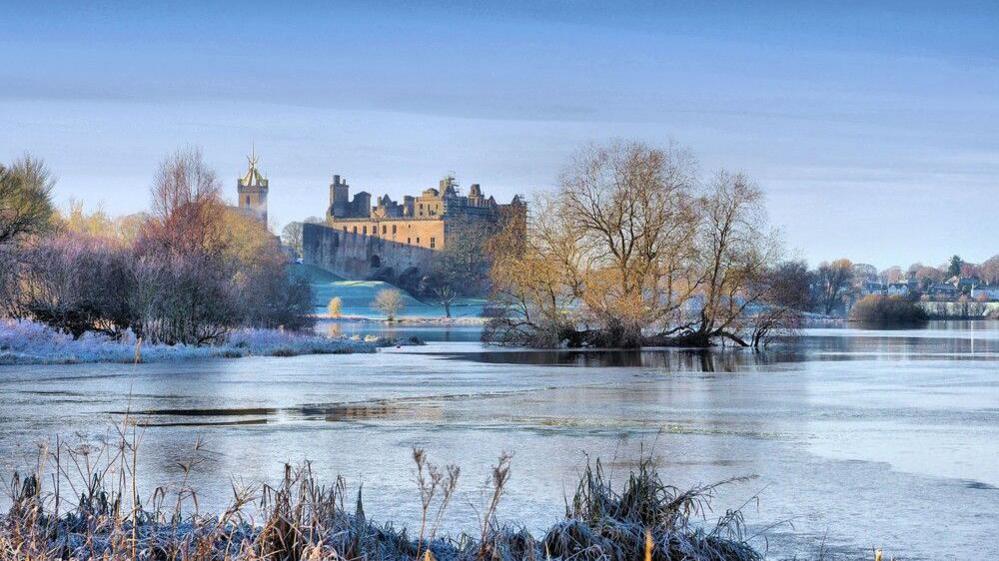 A partially frozen lake by Linlithgow Palace with a blue sky, trees and winter sun.