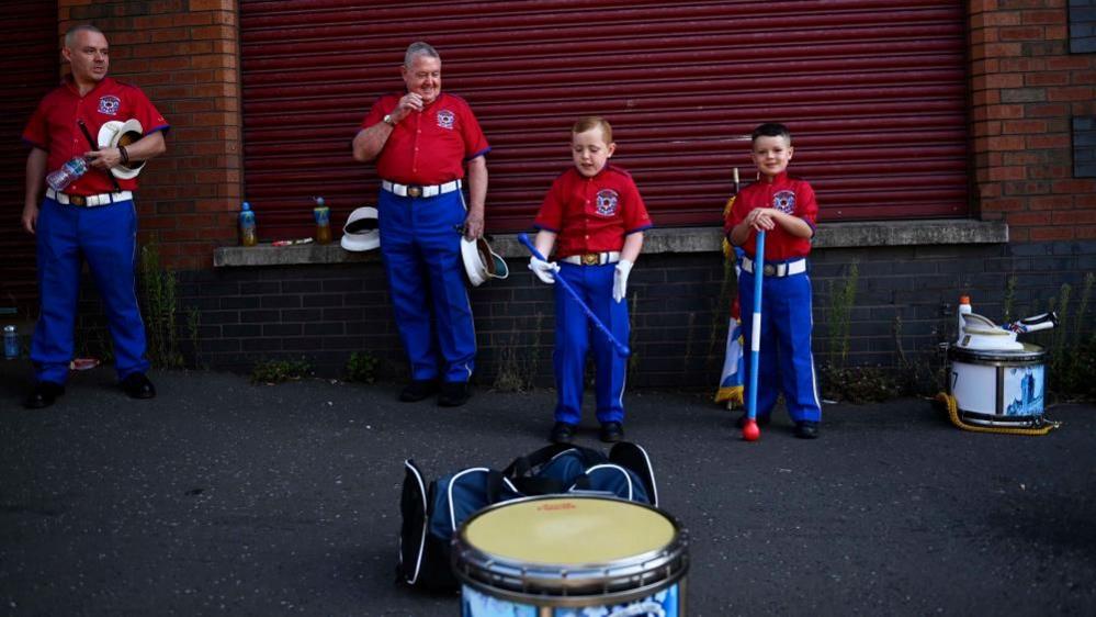 Members of the orange Order parades wait on Clifton Street dressed in red tops, blue trousers and surrounded by drums.