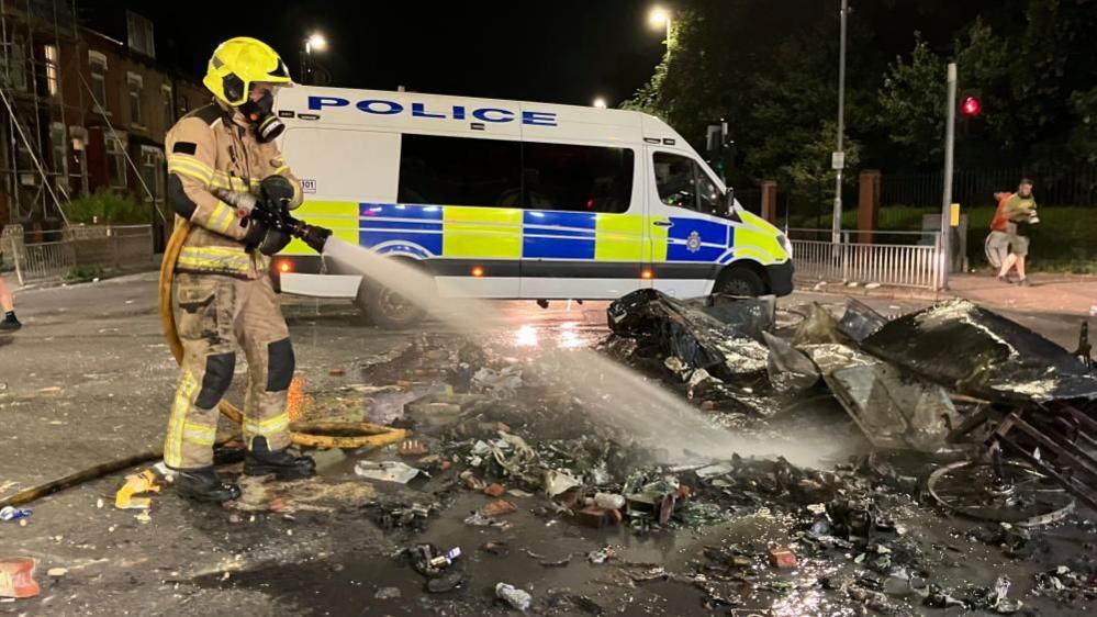A firefighter sprays water at a burnt out vehicle. Behind him is a police van. It is dark and it is a residential street.