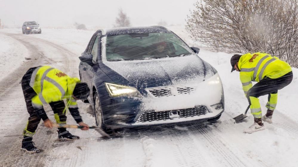 Two men in high vis clothing use spades to dig a blue car out of the now