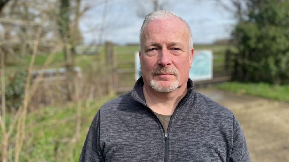 Richard Fuller looks directly at the camera. He has grey hair, a goatee beard and is wearing a grey zip-up top over an olive green T-shirt. He is standing outside with trees and a gate visible behind him.