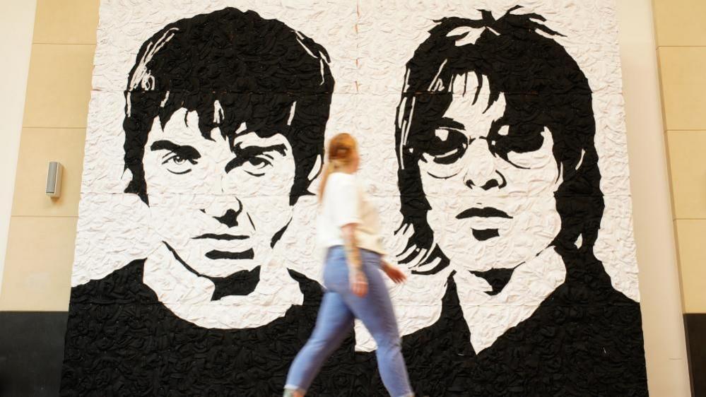 A woman walks past a large black and white mural of Noel and Liam Gallagher made out of bucket hats.