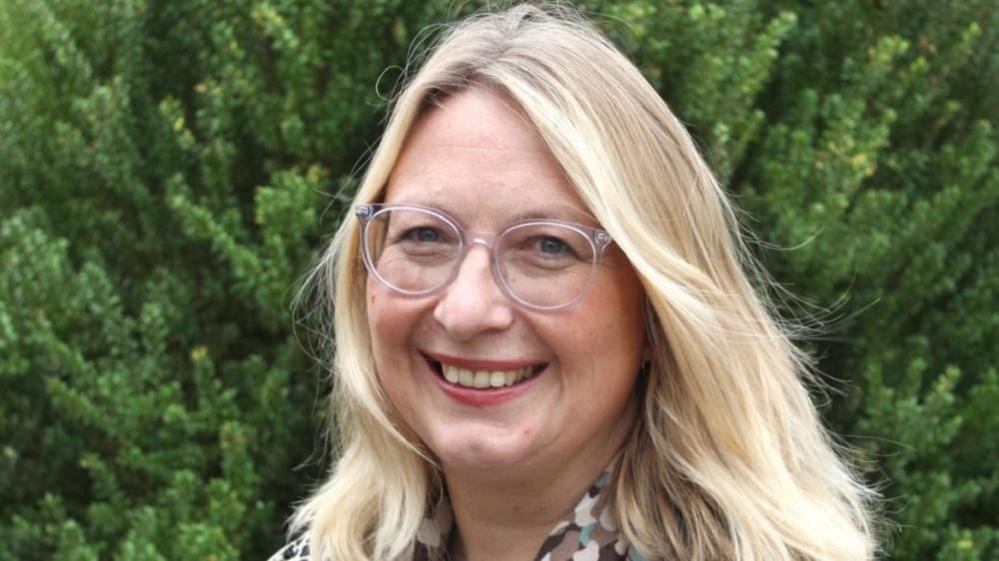 Judi Newman has long blonde hair and is wearing clear framed rounded glasses. She is smiling, and standing near a fir hedge. Part of her blouse, white, patterend with beige, blue and brown shapes, is partly visible.