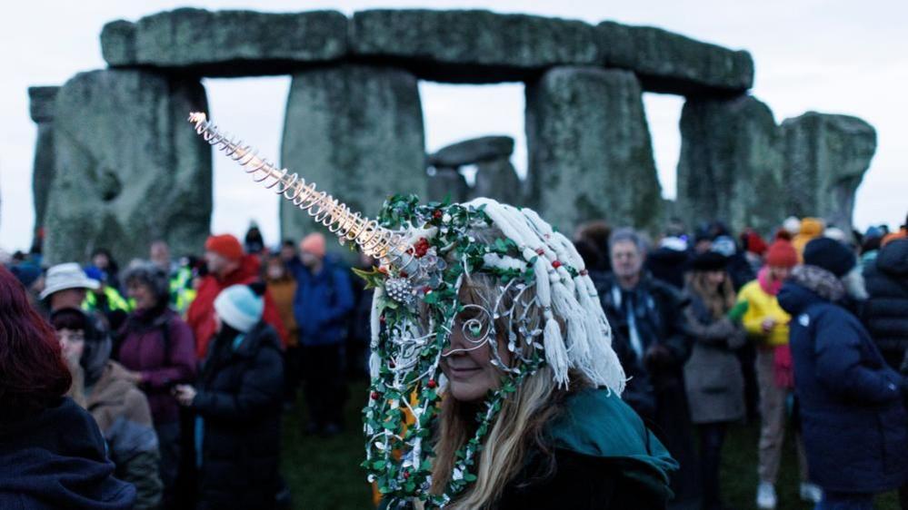 A woman wearing a colourful unicorn hat and mask in front of Stonehenge