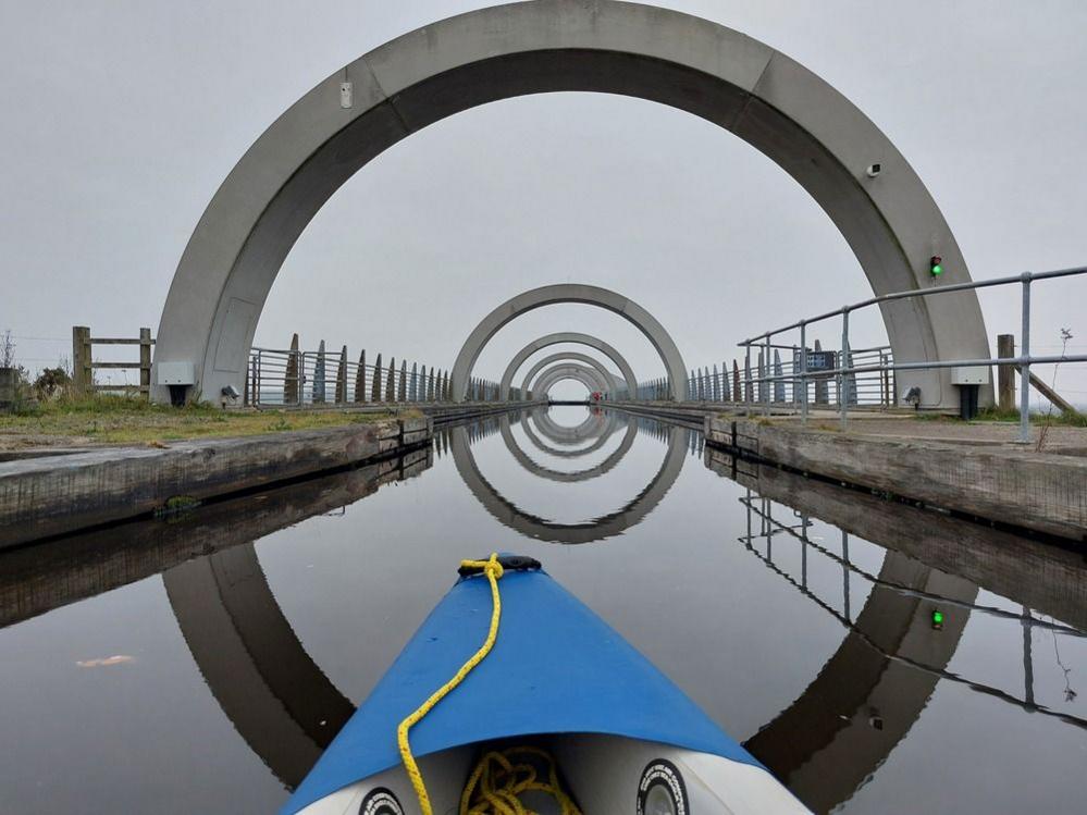 It's a very cloudy and misty day. A view of the Falkirk wheel from a kayak on the waterway. You can see one end of the kayak and five steel arches over the calm waterway.