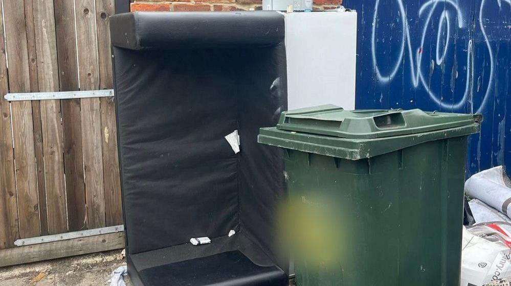 Part of a black sofa without its cushions standing up on its side beside a green general waste bin with the Newcastle City Council logo on it. There's graffiti on the blue wooden door behind the sofa and a pile of rubbish can be seen behind the bin to the right. The number on the bin has been blurred out.