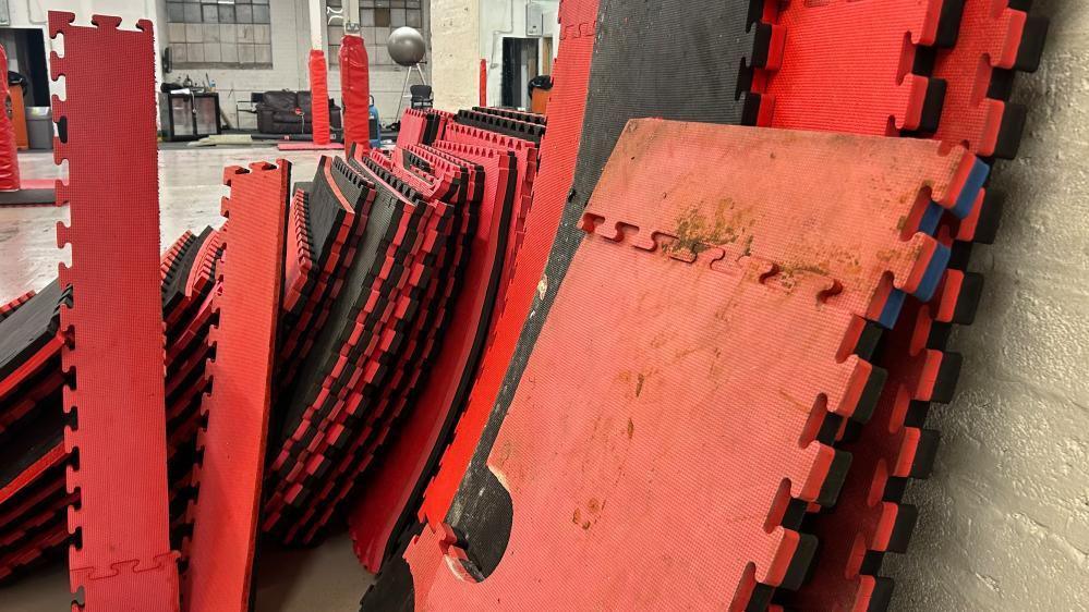 Red and black martial arts training mats piled up against the wall, covered in dirt and sewage after flooding.