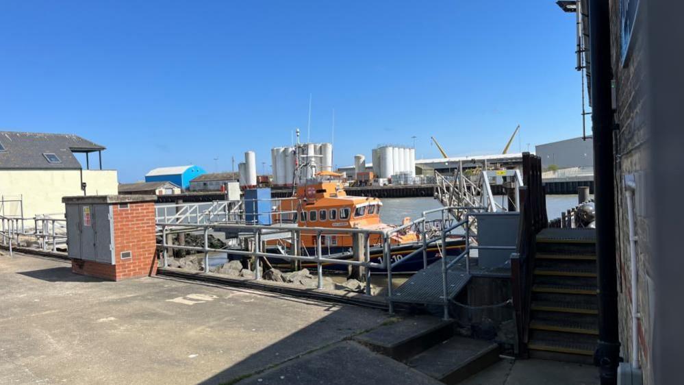 The Gorleston Trent class lifeboat in its mooring at Gorleston Lifeboat Station. It is framed by handrails and a drawbridge for access. In the foreground is concrete hardstanding, and a set of steps up to the lifeboat station. Beyond the River Yare is a set of storage silos and other port buildings in Great Yarmouth.