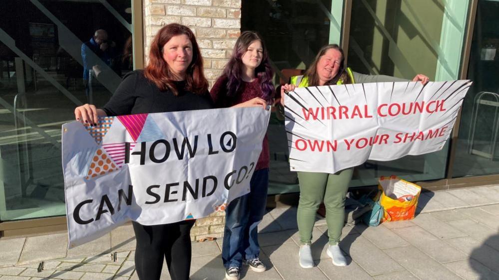 Two women and a girl stand side by side. The two women are holding two different signs which read, "how lo can send go?" and "Wirral Council own your shame". 