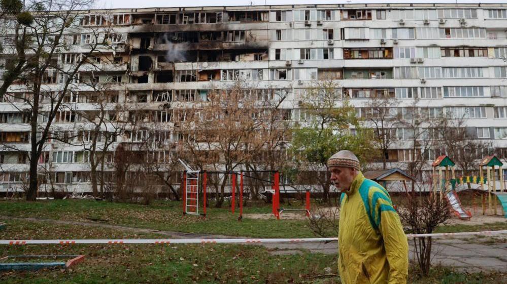 A man in a yellow jacket walking past an apartment block with visible damage from a strike