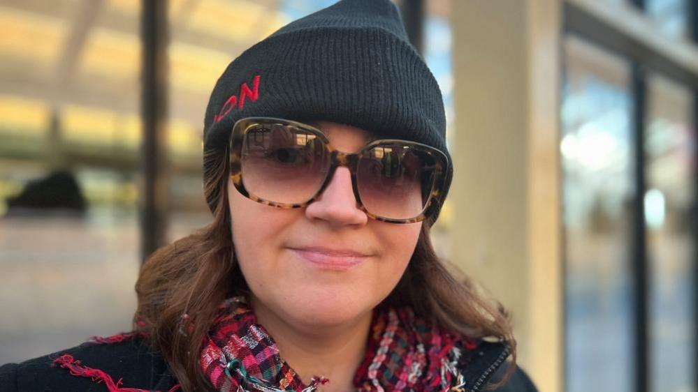 Alexandra Meichan, wearing dark sunglasses, a dark hat, a scarf, looking at the camera and smiling 