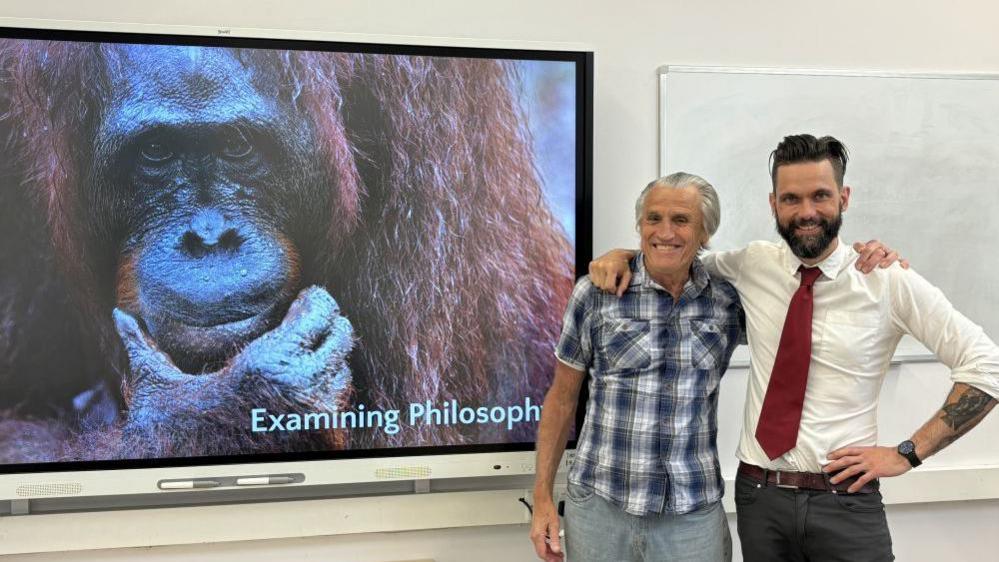 Bob Lakin and Chris Higginson stood with one another in a classroom. Behind them is a whiteboard with an image of an orangutan on and the words "examining philosophy".