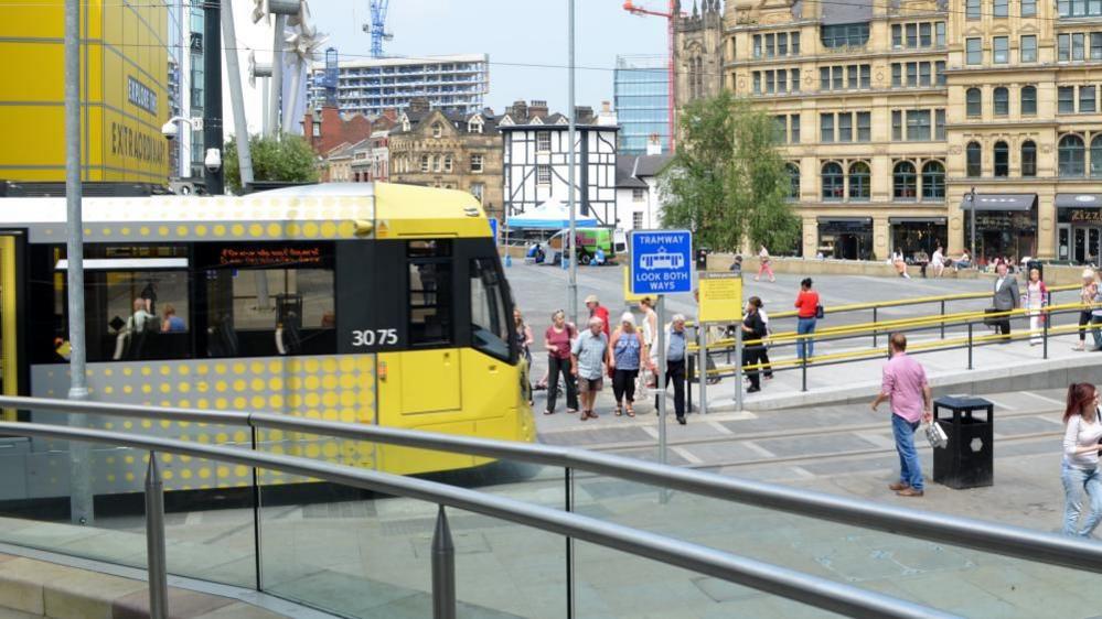 A yellow tram travels along a tram link in Manchester city centre. A number of people are milling and walking around.