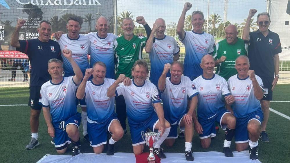 The England over-60s team posing for a photo in two rows. They are wearing blue and white kit. They are posing with the trophy and an England flag.