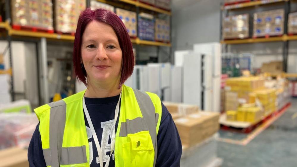 A woman with a purple bob wears a high-vis vest and stands in a warehouse. 